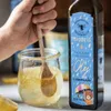 A hand is stirring a jar of honey with a wooden spoon, next to a bottle of oil with a colorful label. Produktbild-Vorschau