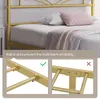A close-up view of a gold metal bed frame, showing details of the built-in slats and how they prevent the mattress from sliding. The top part of the image shows the headboard and a portion of the mattress with pillows and a blanket. Produktbild-Vorschau