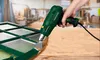 A person is using a green heat gun to strip paint from a green window frame. The window frame is on a wooden surface. Produktbild-Vorschau