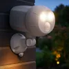 A white motion-activated outdoor spotlight mounted on a brick wall at dusk. Produktbild-Vorschau