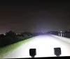 Two black LED work lights are mounted on the front of a vehicle, illuminating a dark road at night. The lights are bright and cast a wide beam, creating a strong glare on the road surface. The surrounding environment is dark, with trees and distant lights  Produktbild-Vorschau