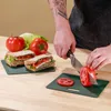 A person is preparing sandwiches and slicing tomatoes on a wooden table. Produktbild-Vorschau