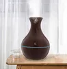 A dark brown vase-shaped aroma diffuser with a blue light strip and a power button symbol is placed on a wooden table next to a glass of water. White curtains are visible in the background. Produktbild-Vorschau