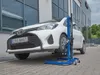 A white Toyota Yaris car is being lifted by a blue hydraulic jack in front of a modern building. Produktbild-Vorschau
