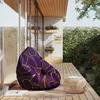 A purple bean bag with a gold floral pattern sits on a wooden deck next to a small woven stool. The scene is set outdoors with lush greenery and a stone wall in the background. Náhled obrázku produktu