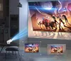 A projector is shown projecting a movie onto a wall in a dimly lit room. The projector is small and white, placed on a table next to a stool. The projected image is a colorful scene from a movie. Below the main projection, two smaller images show the proje Náhled obrázku produktu