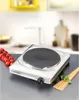 A silver electric hot plate with a black burner is shown on a countertop, with cherry tomatoes and pasta in the foreground. The background shows a blurred kitchen scene. Náhled obrázku produktu