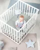 A baby is playing in a white crib with a green mat and a teddy bear. Náhled obrázku produktu