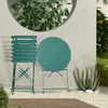 A dark green metal bistro set, consisting of a round table and a chair, is placed on a gravel patio next to a concrete wall with a circular opening revealing greenery. An aloe plant is to the right of the table. Produktbild-Vorschau