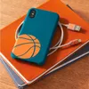 A teal phone case with an orange basketball design is placed on top of two notebooks, with a charging cable connected to the phone. Náhľad obrázku produktu