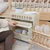 A child's hand is shown opening the door of a wooden toy dishwasher, with dishes in a drying rack nearby. The scene is set in a nursery or playroom. Voir les images du produit