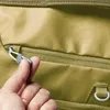 A close-up shot of a hand holding a zipper pull on a khaki-colored bag. Produktbild-Vorschau