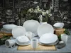 A collection of white ceramic dinnerware, including plates, bowls, and mugs, arranged on a table with a chalkboard-style background featuring various text and illustrations. Produktbild-Vorschau