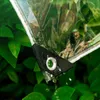 A close-up of a clear PVC tarp with water dripping from a grommet, set against a background of green leaves. Produktbild-Vorschau