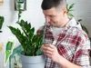 A man is tending to a potted plant, holding a small green dropper near its leaves. There is a separate image of a green dropper with text on it. Produktbild-Vorschau