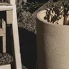 A close-up view of a textured cream-colored planter with a plant inside, next to a wooden chair leg. Produktbild-Vorschau