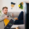 A man is relaxing on a couch, holding a remote control and a mug, with a plate of croissants nearby. A television remote is prominently displayed in the foreground. Produktbild-Vorschau