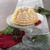 A bundt cake shaped like a pine forest is dusted with powdered sugar and displayed on a glass cake stand, surrounded by festive holiday decorations like pinecones and red berries. Anteprima dell’immagine prodotto