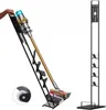 A hand is holding a Dyson vacuum cleaner, which is placed on a black stand with various attachments. Another black stand is shown separately on the right. Produktbild-Vorschau