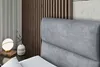 A close-up view of a bed with a gray upholstered headboard and a bedside table with a lamp and vase. Produktbild-Vorschau