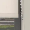 A close-up view of a beige roller blind with a beaded pull cord and a clear plastic cord holder, seen from the side, with a blurred outdoor scene visible through a window. Produktbild-Vorschau