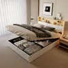 A bed with its storage compartment open, revealing stored items. The bed is in a bedroom setting with curtains and a bedside table visible. Produktbild-Vorschau