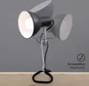A black metal clamp lamp with a silver articulated arm is attached to a wooden surface. The lamp is illuminated, and a ghosted image of the lamp in a different position is visible behind it. Produktbild-Vorschau