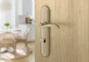 A close-up of a bronze door handle on a wooden door, with a blurred background of a dining area. Produktbild-Vorschau
