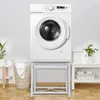 A white washing machine is placed on a white metal stand in a room with white walls and a wooden floor. There are laundry baskets and shelves with towels and detergent bottles visible. Produktbild-Vorschau
