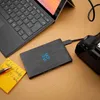 A black external hard drive is placed on a brown surface next to a laptop and a camera. Produktbild-Vorschau