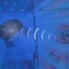 A baby projector with a whale design is projecting a starry sky and fish onto a crib, with a baby lying in the crib. Náhled obrázku produktu