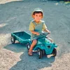 A young child is riding a teal toy tractor with a trailer in an outdoor setting. Produktbild-Vorschau
