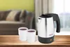 A travel kettle and two cups are placed on a wooden table in front of a blurred living room background. Produktbild-Vorschau