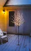 A decorative LED tree with white lights stands on a snow-covered deck next to a chair, with a house in the background. Produktbild-Vorschau