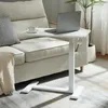 A white adjustable laptop table is placed next to a sofa, with a laptop and a cup of coffee on it. Náhľad obrázku produktu