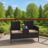 A black rattan bench with cream cushions and a small table sits on a wooden deck, with a lush green forest and blue sky in the background. Produktbild-Vorschau