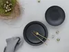A top-down view of a table setting with dark plates, cutlery, a nest of quail eggs, and a grey napkin. Produktbild-Vorschau
