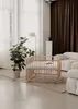 A wooden playpen is set up in a bright, modern living room with large windows and sheer curtains. A large potted plant is nearby, and a plush white sofa is visible in the background. Produktbild-Vorschau