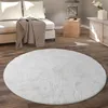 A round, fluffy white rug is placed on a wooden floor in a living room setting, with a sofa and side table in the background. Produktbild-Vorschau