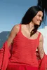 A woman wearing a red knitted top and cardigan, smiling outdoors. Produktbild-Vorschau