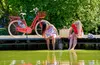 Two women sitting on a wooden pier with their feet in the water, next to a red bicycle. Produktbild-Vorschau
