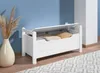 A white storage bench with its lid open, revealing pillows and blankets inside, placed in a room with a blue wall and a patterned rug. Produktbild-Vorschau