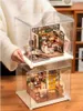 Two miniature dollhouse kits are displayed inside clear plastic cases, stacked on top of each other. The top kit appears to be a coffee shop, while the bottom kit is a kitchen scene. A person's hands are visible holding the top case. Podgląd zdjęcia produktu