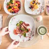 A table setting with a variety of dishes and food items, including steak, salad, fruit, and dessert, arranged on plates with a floral design. Voir les images du produit