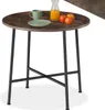 A round dining table with a dark wood-effect top and black metal legs, set against a white background. There are a few items on the table: a green mug, a green plate, a glass jug with orange juice, and three small fruits (two red, one orange). Produktbild-Vorschau