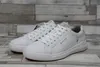 A pair of white leather sneakers are displayed on a wooden surface against a grey and white wood-paneled background. Náhľad obrázku produktu