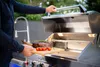 A person is holding a cutting board with a steak and tomatoes, about to place it on a grill. Podgląd zdjęcia produktu