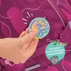A hand is holding a unicorn patch near a backpack. There is also a tag with text and an illustration. Anteprima dell’immagine prodotto