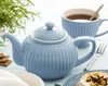 A blue teapot and cup are set on a table with crackers and a piece of cheese, suggesting a tea time setting. Produktbild-Vorschau