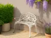 A white metal garden bench with ornate floral detailing sits on a paved patio, surrounded by lush green plants and purple wisteria flowers. Náhľad obrázku produktu